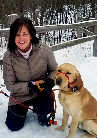 woman smiling at camera and kneeling on snow next to yellow Lab