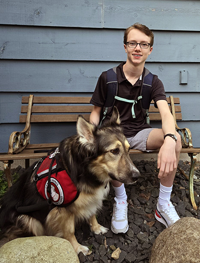 Teenage boy sitting on bench outside, smiling at camera, with hand on head of German Shepherd mix dog wearing red service cape