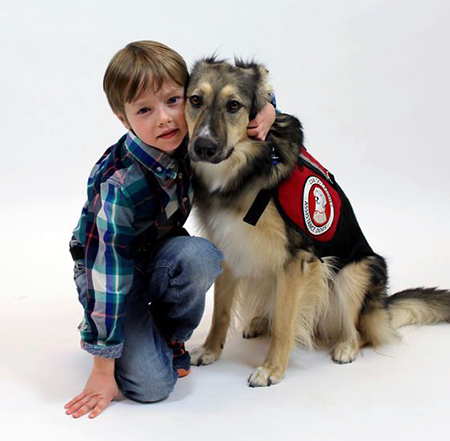 Boy kneeling on floor with arm around and cheek against German Shepherd mix dog wearing red service cape; both are looking at camera