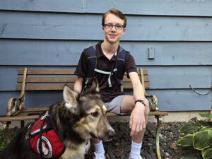 Teenage boy sitting on bench outside, smiling at camera, with hand on head of German Shepherd mix dog wearing red service cape