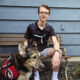 Teenage boy sitting on bench outside, smiling at camera, with hand on head of German Shepherd mix dog wearing red service cape