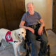 man sitting in chair in home with yellow Lab service dog wearing red cape standing at his side