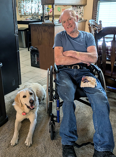 man sitting in a wheelchair in home with yellow Lab service dog wearing red cape lying at his side