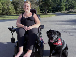 woman sitting in wheelchair on street with black Lab service dog wearing red cape sitting next to her; both are smiling at the camera