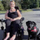 woman sitting in wheelchair on street with black Lab service dog wearing red cape sitting next to her; both are smiling at the camera