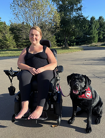 woman sitting in wheelchair on street with black Lab service dog wearing red cape sitting next to her; both are smiling at the camera