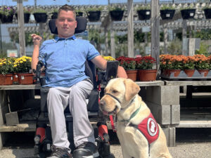 young man sitting in power chair in front of flowering plants with yellow Lab service dog wearing red cape sitting next to him