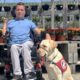 young man sitting in power chair in front of flowering plants with yellow Lab service dog wearing red cape sitting next to him