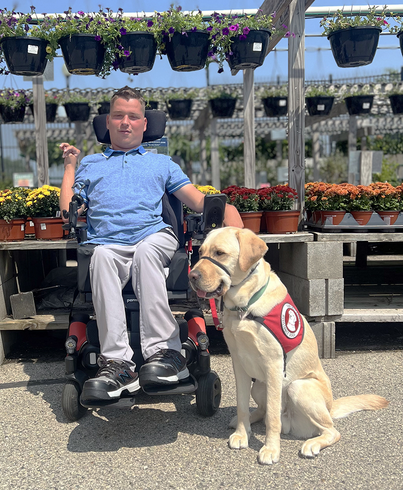 young man sitting in power chair in front of flowering plants with yellow Lab service dog wearing red cape sitting next to him