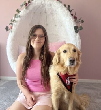 girl sitting on bedroom carpet with hand on Golden Retriever that is wearing a red service cape; both are looking at the camera