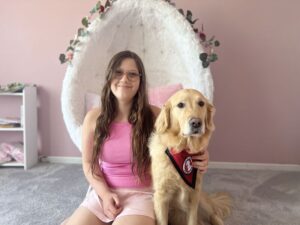 girl sitting on bedroom carpet with hand on Golden Retriever that is wearing a red service cape; both are looking at the camera