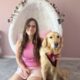 girl sitting on bedroom carpet with hand on Golden Retriever that is wearing a red service cape; both are looking at the camera