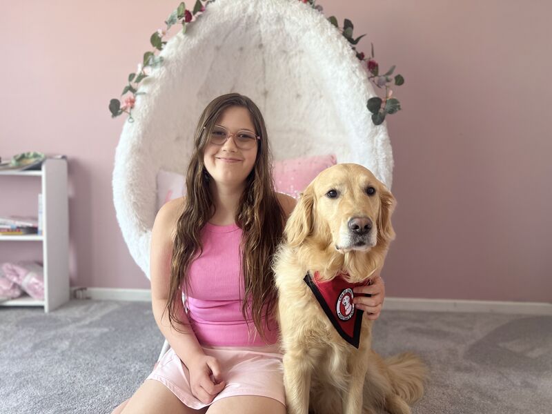 girl sitting on bedroom carpet with hand on Golden Retriever that is wearing a red service cape; both are looking at the camera