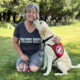 woman kneeling on grass and smiling at camera while yellow Lab dog wearing red Can Do Canines cape licks her chin