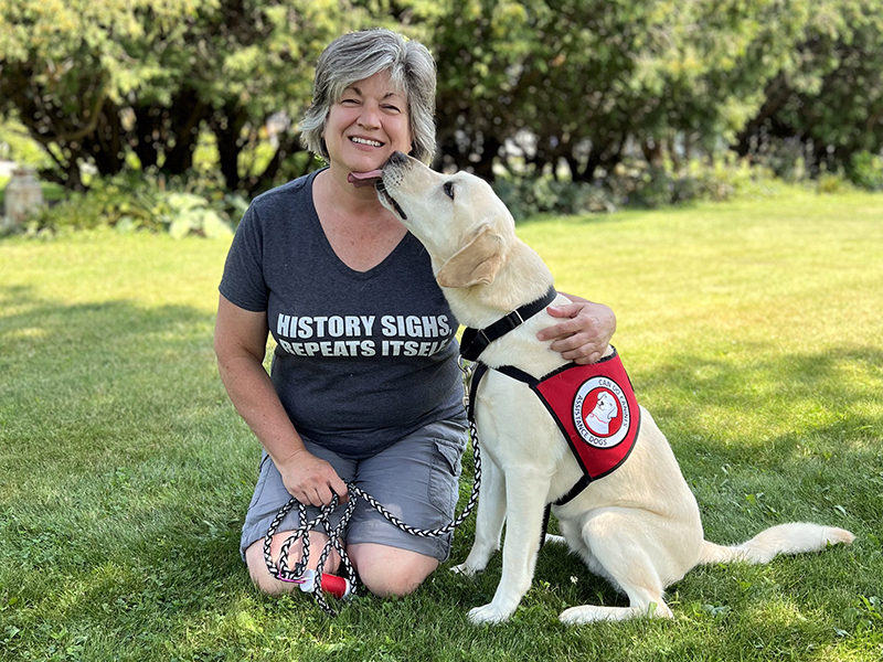 woman kneeling on grass and smiling at camera while yellow Lab dog wearing red Can Do Canines cape licks her chin