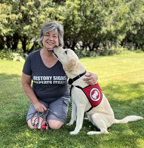 woman kneeling on grass and smiling at camera while yellow Lab dog wearing red Can Do Canines cape licks her chin