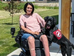 woman sitting outside in wheelchair with black Lab service dog wearing red Can Do Canines cape with front paws on her leg; both are smiling at camera