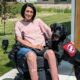 woman sitting outside in wheelchair with black Lab service dog wearing red Can Do Canines cape with front paws on her leg; both are smiling at camera