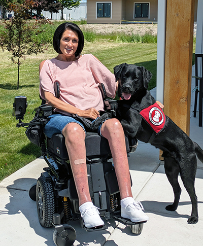 woman sitting outside in wheelchair with black Lab service dog wearing red Can Do Canines cape with front paws on her leg; both are smiling at camera