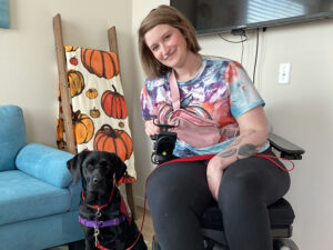 woman sitting in wheelchair and leaning head down toward black Lab dog sitting next to her; both are smiling at camera