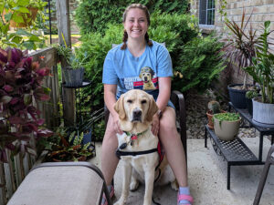 young woman sitting on patio chair outside with a yellow Lab dog sitting between her knees; both are smiling at camera
