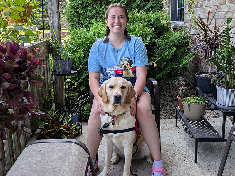 young woman sitting on patio chair outside with a yellow Lab dog sitting between her knees; both are smiling at camera