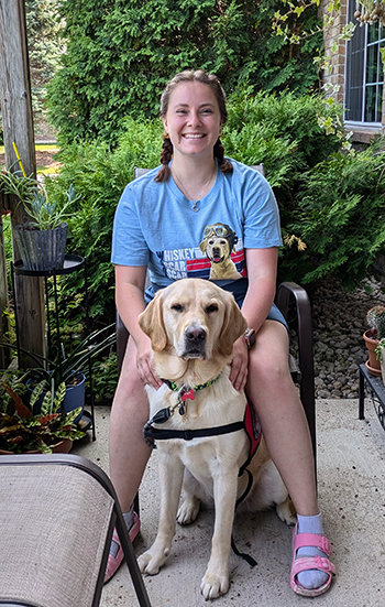 young woman sitting on patio chair outside with a yellow Lab dog sitting between her knees; both are smiling at camera