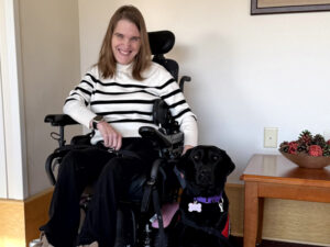 young woman sitting indoors in a wheelchair with hand on black Lab sitting next to her
