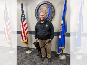 Police officer standing in front of Coon Rapids Police Department wall seal and between two flags with black Lab service dog sitting next to him