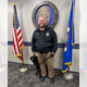Police officer standing in front of Coon Rapids Police Department wall seal and between two flags with black Lab service dog sitting next to him