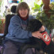 woman sitting in wheelchair in home with black Lab dog wearing red Can Do Canines service cape at her side, putting front paws on her lap; both are smiling at camera