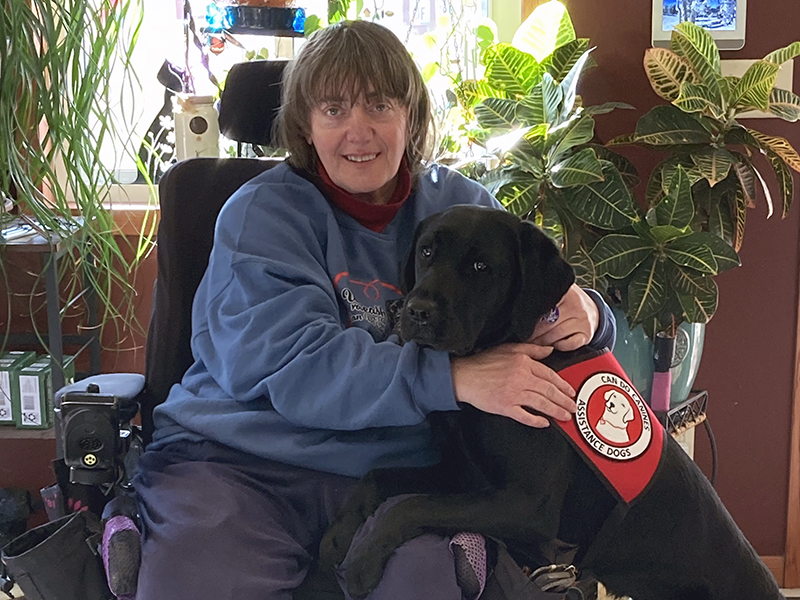 woman sitting in wheelchair in home with black Lab dog wearing red Can Do Canines service cape at her side, putting front paws on her lap; both are smiling at camera