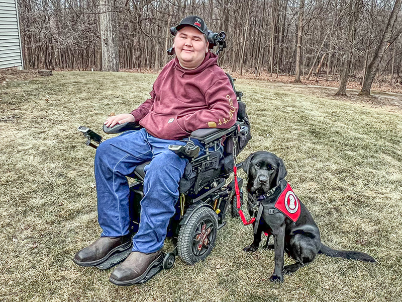 young man sitting outside in wheelchair with black Lab wearing red service cape sitting beside him