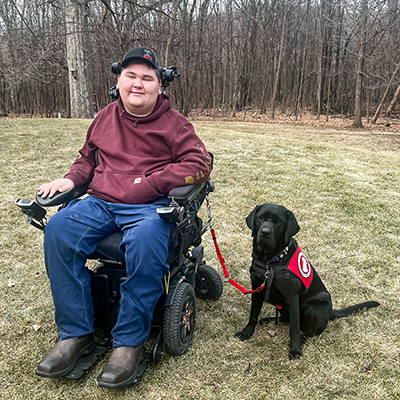 young man sitting outside in wheelchair with black Lab wearing red service cape sitting beside him