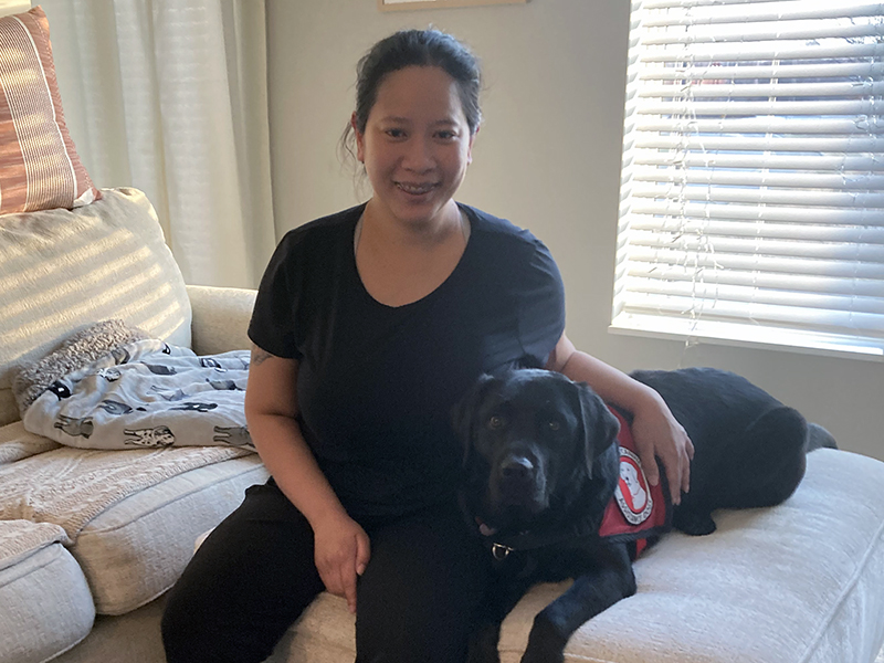 woman sitting on ottoman with arm around black Lab dog wearing red service cape; both are smiling at camera
