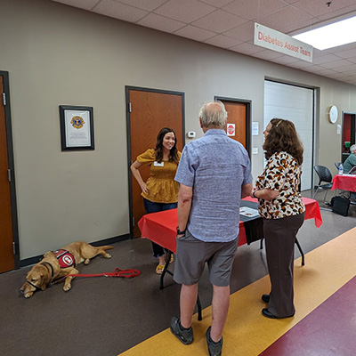 woman standing at table, talking with couple on other side, with service dog lying near her