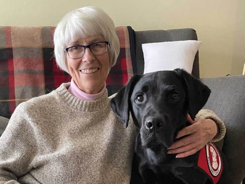 A woman sits on the floor with her arm around her black lab