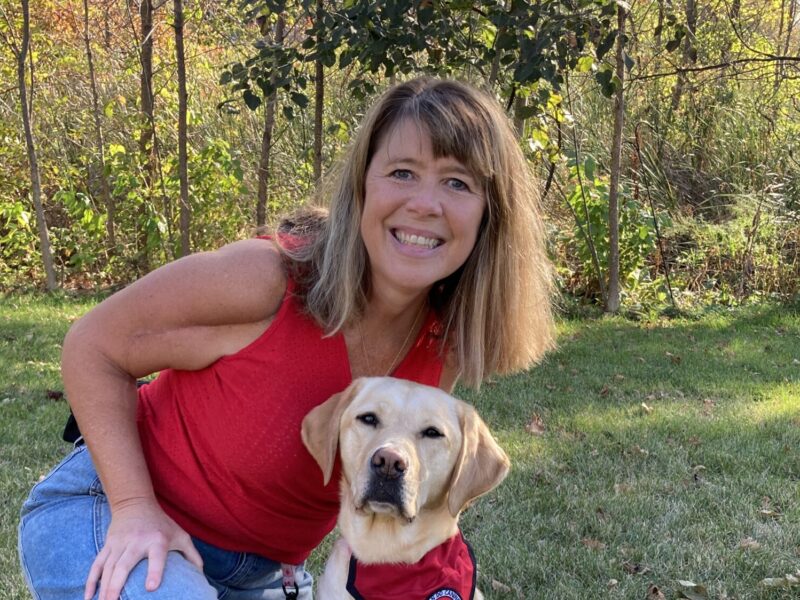 A smiling woman sits with her arm around her yellow lab