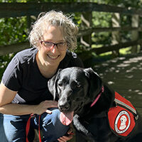 woman crouching on walking bridge with hand on black Lab wearing red Can Do Canines service cape; both are smiling at camera