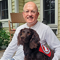 man sitting on front porch with arm around brown Spaniel wearing red service vest; both are smiling at the camera