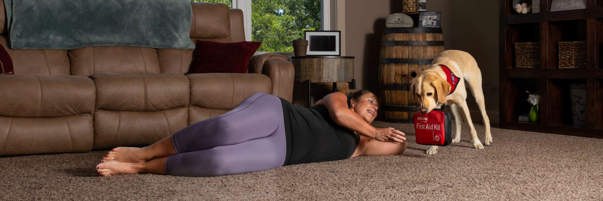 woman lying on her side on living room floor with her hand outstretched toward a yellow Lab service dog wearing Can Do Canines cape bringing her a first aid bag